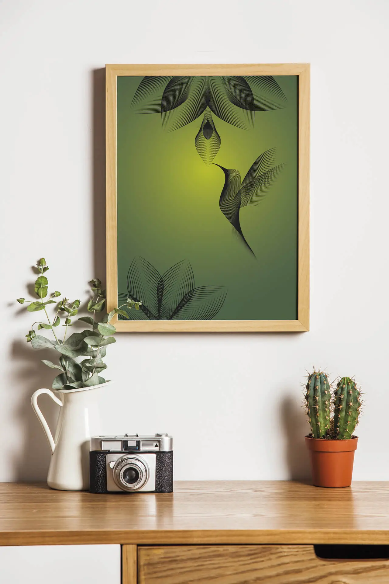 Illustration de colibri encadrée accrochée à un mur au dessus d'une composition faite de plante et d'un appareil photo rétro